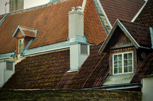 The image features a series of steeply pitched roofs with red clay tiles and wooden accents. There is a prominent white chimney, flanked by weathered roofing. A window with detailed wood framing is visible, hinting at a rustic architectural style.