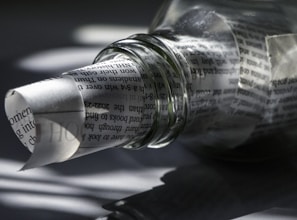 A transparent glass bottle lies on its side, partially filled with rolled newspaper. The text on the paper is visible through the bottle's clear surface, creating a distorted, artistic effect. Shadows and light play across the surface, adding depth and intrigue.