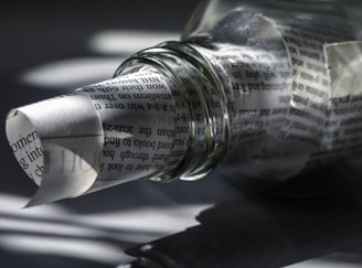 A transparent glass bottle lies on its side, partially filled with rolled newspaper. The text on the paper is visible through the bottle's clear surface, creating a distorted, artistic effect. Shadows and light play across the surface, adding depth and intrigue.