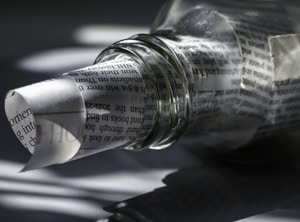 A transparent glass bottle lies on its side, partially filled with rolled newspaper. The text on the paper is visible through the bottle's clear surface, creating a distorted, artistic effect. Shadows and light play across the surface, adding depth and intrigue.