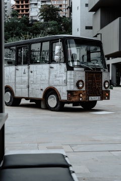 A vintage-styled vehicle with metal paneling parked in an urban setting. The vehicle has rectangular windows and round headlights, and its body is decorated with a pattern resembling soldered metal plates. It is situated in a modern area with buildings in the background and greenery further back.