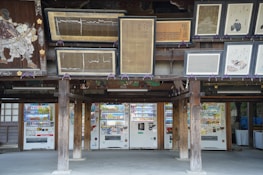 An arrangement of traditional Japanese artwork adorns the upper section of a wooden structure, beneath which are several modern vending machines. The combination of classic art and contemporary technology creates an interesting juxtaposition.
