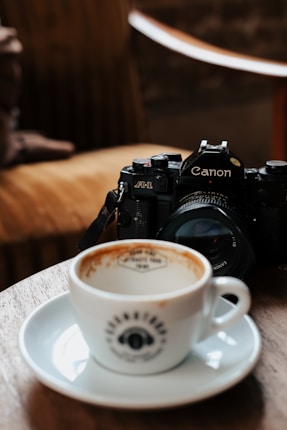 A vintage Canon A-1 camera rests on a wooden surface next to a white coffee cup with a saucer. The cup contains remnants of coffee, suggesting it's been partially consumed. The image is set in a cozy interior with warm lighting and a blurred background, featuring what appears to be a chair, creating a relaxed atmosphere.