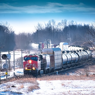 A freight train travels through a snowy landscape, with its bright red engine leading a series of silver tank cars. Trees with bare branches line the background, under a clear blue sky with some wispy clouds.