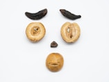 Various nuts and dried fruits arranged on a white surface to resemble a face, with two long dark fruits as eyebrows, two round nuts as eyes, a small dark nut as the nose, and a larger nut as the mouth.