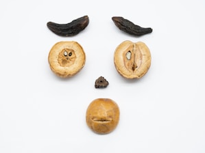 Various nuts and dried fruits arranged on a white surface to resemble a face, with two long dark fruits as eyebrows, two round nuts as eyes, a small dark nut as the nose, and a larger nut as the mouth.