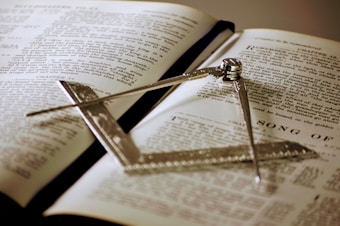 An open book displaying text in English, with a metallic square and compass placed on top. The compass is positioned diagonally across the pages.