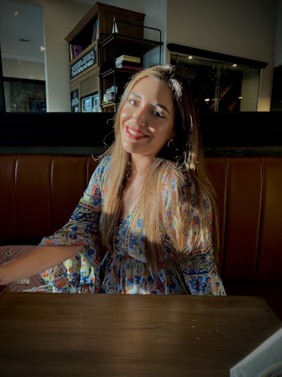A woman with long, straight hair is sitting in a warmly lit setting. She wears a colorful, floral-patterned dress and smiles brightly towards the camera. The sunlight casts a gentle glow on her face and hair, creating a relaxed and inviting atmosphere. In the background, there are shelves with books and wine racks.