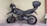 A black motorcycle with a top case is parked in a corner, resting on its kickstand. The motorcycle has rugged tires suitable for off-road use, and a helmet is placed on the handlebar. The background consists of a plain, light-colored wall, and the ground is marked with parking lines.