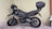A black motorcycle with a top case is parked in a corner, resting on its kickstand. The motorcycle has rugged tires suitable for off-road use, and a helmet is placed on the handlebar. The background consists of a plain, light-colored wall, and the ground is marked with parking lines.