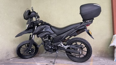 A black motorcycle with a top case is parked in a corner, resting on its kickstand. The motorcycle has rugged tires suitable for off-road use, and a helmet is placed on the handlebar. The background consists of a plain, light-colored wall, and the ground is marked with parking lines.