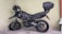 A black motorcycle with a top case is parked in a corner, resting on its kickstand. The motorcycle has rugged tires suitable for off-road use, and a helmet is placed on the handlebar. The background consists of a plain, light-colored wall, and the ground is marked with parking lines.