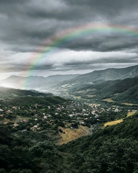 A scenic valley with lush green hills and a village nestled between them. A vibrant rainbow arches across the sky against a backdrop of dramatic, cloudy skies.