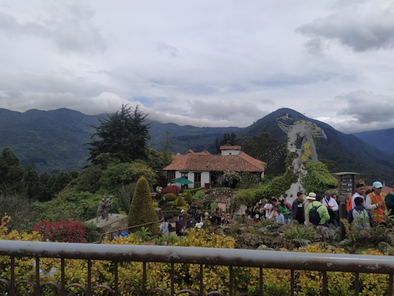 A scenic mountain landscape featuring a rustic house with a red-tiled roof surrounded by lush greenery and colorful shrubs. A group of people is gathered in the foreground, near a large decorative sculpture covered in foliage. The background is dominated by rolling mountains under an overcast sky.