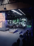 A large indoor venue with high ceilings and industrial architecture. Rows of chairs are arranged facing a large screen on the left. A gathering of people is seated, while others stand at the back and sides. The atmosphere is dimly lit with green lighting accentuating some areas, including small trees near the entrance. The ceiling features metal beams with lights suspended from them.