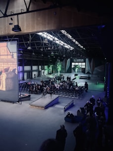 A large indoor venue with high ceilings and industrial architecture. Rows of chairs are arranged facing a large screen on the left. A gathering of people is seated, while others stand at the back and sides. The atmosphere is dimly lit with green lighting accentuating some areas, including small trees near the entrance. The ceiling features metal beams with lights suspended from them.