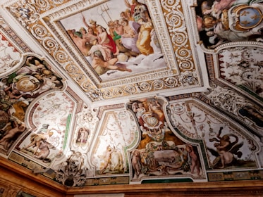 Elaborate ceiling artwork featuring multiple mythological scenes bordered with intricate gold and white detailing. Central panel depicts a group of deities, possibly from Greek mythology, in various poses, surrounded by smaller scenes showing figures, some with wings or in dramatic poses, and decorative elements such as cherubs and scrolls.