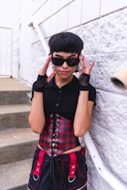 A person with short hair wearing a black top and plaid red corset stands on concrete steps, posing with hands on the sides of bold, dark sunglasses. The outfit includes fingerless gloves and pants with red accents against a textured white wall backdrop.
