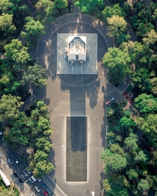 Aerial view of a large rectangular monument surrounded by green trees, with a pathway leading up to it and a rectangular plaza area in front. The monument appears to be in a circular paved area within a park-like setting. There are several cars visible on the road beside the park.