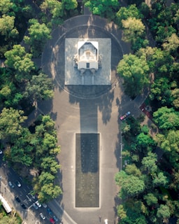 Aerial view of a large rectangular monument surrounded by green trees, with a pathway leading up to it and a rectangular plaza area in front. The monument appears to be in a circular paved area within a park-like setting. There are several cars visible on the road beside the park.