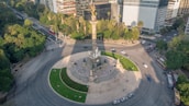 An aerial view of a large, roundabout with a monument in the center, surrounded by green trees and busy city streets. The monument features a tall column topped with a golden figure. The area is bordered by modern office buildings, and several vehicles are visible on the roads. Pedestrians are scattered around the circular plaza, which has neatly maintained green patches.