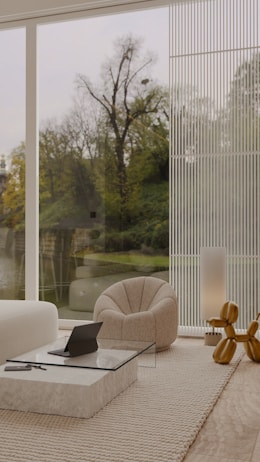 A modern and minimalist interior living space with a large floor-to-ceiling window offering a view of a natural landscape with trees and a body of water. The room features a comfortable textured armchair, a low glass-topped coffee table on a beige rug, and a sleek sofa. A decorative object resembling golden balloons and a tall lamp add a contemporary touch. A laptop sits on the coffee table.