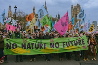 A large group of people are gathered in a city street, participating in a protest or demonstration. They are holding a green banner with the words 'NO NATURE NO FUTURE' written on it. Various colorful flags with images of animals and slogans are being waved by the attendees. The backdrop includes historical architecture, suggesting a significant urban location.