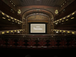 A grand, ornate theater with a large screen at the center, surrounded by elegantly designed architectural elements. The interior features intricate patterns and warm lighting, with multiple levels of seating and decorative balconies. The atmosphere suggests a historic or classic design theme.