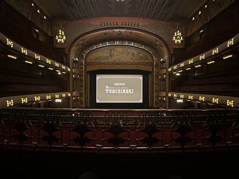 A grand, ornate theater with a large screen at the center, surrounded by elegantly designed architectural elements. The interior features intricate patterns and warm lighting, with multiple levels of seating and decorative balconies. The atmosphere suggests a historic or classic design theme.