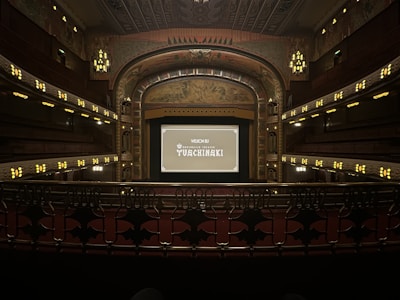 A grand, ornate theater with a large screen at the center, surrounded by elegantly designed architectural elements. The interior features intricate patterns and warm lighting, with multiple levels of seating and decorative balconies. The atmosphere suggests a historic or classic design theme.