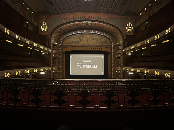 A grand, ornate theater with a large screen at the center, surrounded by elegantly designed architectural elements. The interior features intricate patterns and warm lighting, with multiple levels of seating and decorative balconies. The atmosphere suggests a historic or classic design theme.