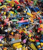 A colorful assortment of keychains and small plastic figurines featuring various cartoon characters and designs. The items are packed closely together, creating a vibrant and chaotic visual effect. Among the collection, there are wristbands, toys, and a variety of character-themed accessories.