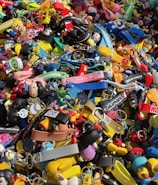 A colorful assortment of keychains and small plastic figurines featuring various cartoon characters and designs. The items are packed closely together, creating a vibrant and chaotic visual effect. Among the collection, there are wristbands, toys, and a variety of character-themed accessories.
