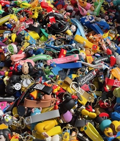 A colorful assortment of keychains and small plastic figurines featuring various cartoon characters and designs. The items are packed closely together, creating a vibrant and chaotic visual effect. Among the collection, there are wristbands, toys, and a variety of character-themed accessories.