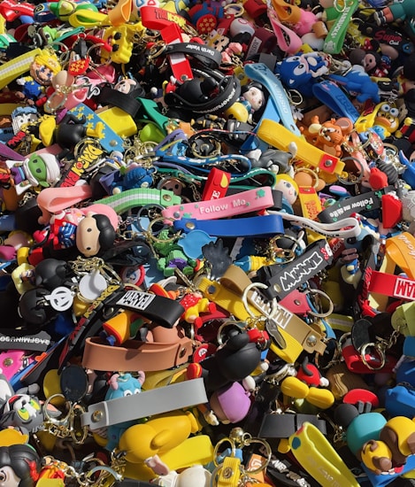 A colorful assortment of keychains and small plastic figurines featuring various cartoon characters and designs. The items are packed closely together, creating a vibrant and chaotic visual effect. Among the collection, there are wristbands, toys, and a variety of character-themed accessories.