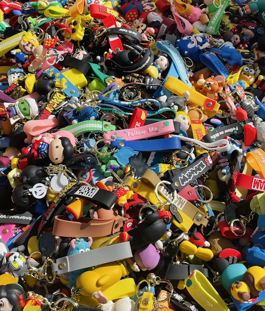 A colorful assortment of keychains and small plastic figurines featuring various cartoon characters and designs. The items are packed closely together, creating a vibrant and chaotic visual effect. Among the collection, there are wristbands, toys, and a variety of character-themed accessories.