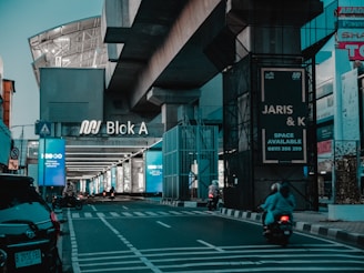 A modern urban setting featuring a large sign that reads 'Blok A' beneath an overpass. Several motorcycles are driving along an empty street flanked by illuminated advertisement panels. Various commercial signs and buildings can be seen in the background.