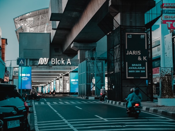 A modern urban setting featuring a large sign that reads 'Blok A' beneath an overpass. Several motorcycles are driving along an empty street flanked by illuminated advertisement panels. Various commercial signs and buildings can be seen in the background.