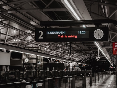 An indoor train station platform featuring a digital sign indicating 'Train is arriving.' The sign includes the location 'Bundaran HI' and the time '18:25,' alongside an analog clock showing the same time. The station has a modern design with exposed metal beams on the ceiling and a series of lights illuminating the platform. Glass barriers line the edge of the platform, and there's a red sign on the right.