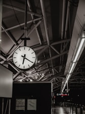 A clock with a black and white face hanging from the ceiling in a modern, industrial-style transport terminal. The architectural features include metal beams and fluorescent lighting, with visible signage indicating train or bus information in the background.