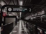 A railway platform with an overhead sign displaying the time and destination. The station appears empty with dim lighting and clean, modern architectural design. The platform number is visible, and the structure includes reflective surfaces and geometric patterns.
