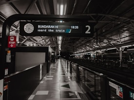 A railway platform with an overhead sign displaying the time and destination. The station appears empty with dim lighting and clean, modern architectural design. The platform number is visible, and the structure includes reflective surfaces and geometric patterns.