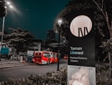 A street scene at night with a red and white articulated bus traveling on a road lined with trees and street lamps. There is a large signboard on the right displaying 'Taman Literasi Martha Christina Tiahahu,' partially illuminated by a streetlight. The atmosphere is serene, with sparse traffic and dim lighting.