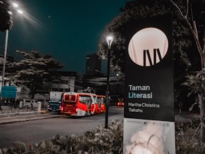 A street scene at night with a red and white articulated bus traveling on a road lined with trees and street lamps. There is a large signboard on the right displaying 'Taman Literasi Martha Christina Tiahahu,' partially illuminated by a streetlight. The atmosphere is serene, with sparse traffic and dim lighting.