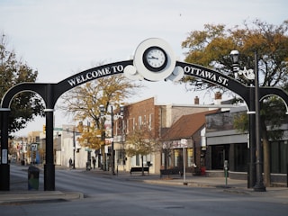 An archway on a street marks the entrance to Ottawa St., featuring a large clock at the center. The street is lined with small businesses and trees, with few people walking around. The area has an inviting and peaceful feel, typical of a small town or community street.