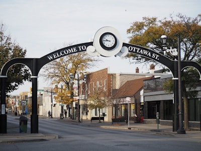 An archway on a street marks the entrance to Ottawa St., featuring a large clock at the center. The street is lined with small businesses and trees, with few people walking around. The area has an inviting and peaceful feel, typical of a small town or community street.