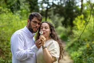 A couple stands in a lush green forest setting, focusing on a dandelion. Both appear to be happily engaged in blowing the seeds into the air, which are visibly floating around them.