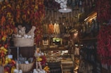 A cozy deli shop filled with a variety of hanging dried sausages and bunches of dried chili peppers. Wicker baskets of garlic and other produce sit on shelves, with an assortment of deli meats and cheeses visible at the back of the shop.