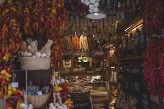A cozy deli shop filled with a variety of hanging dried sausages and bunches of dried chili peppers. Wicker baskets of garlic and other produce sit on shelves, with an assortment of deli meats and cheeses visible at the back of the shop.