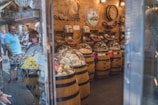 A quaint confectionery store features wooden barrels filled with brightly colored candies, covered with clear dome lids. The barrels are neatly arranged along the store with shelves in the background holding assorted sweets. The warm, wooden interior is adorned with decorative pieces like maps and paintings. Reflections of people observing the display can be seen through the glass exterior.